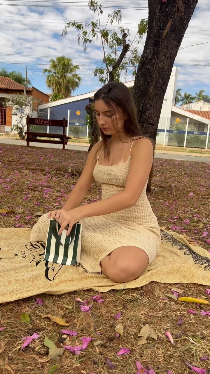 Moça sentada na canga, num parque, abrindo a sacola listrada